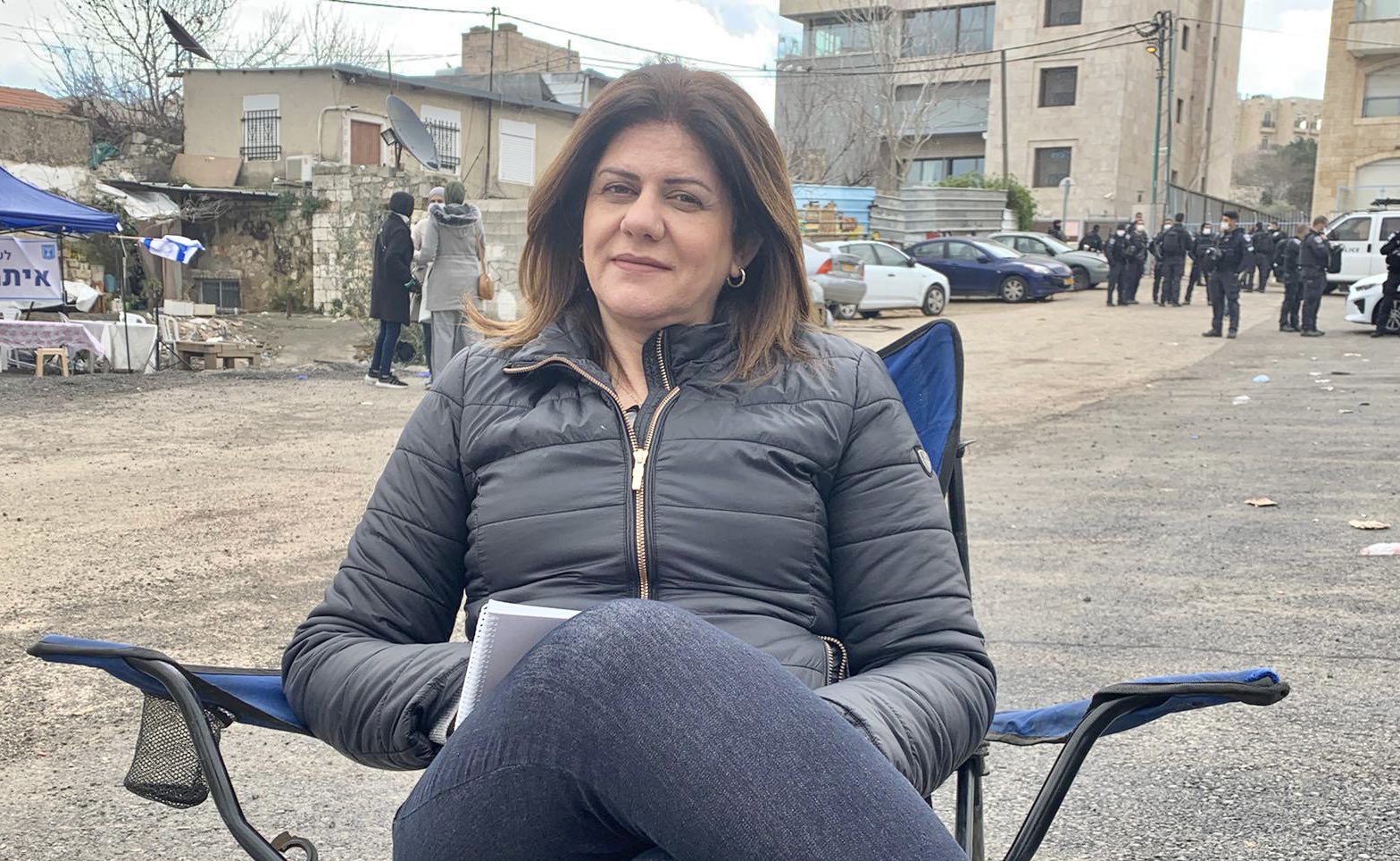 Shireen sits in a chair on the street, smiling wearing a black puffer jacket and jeans. 