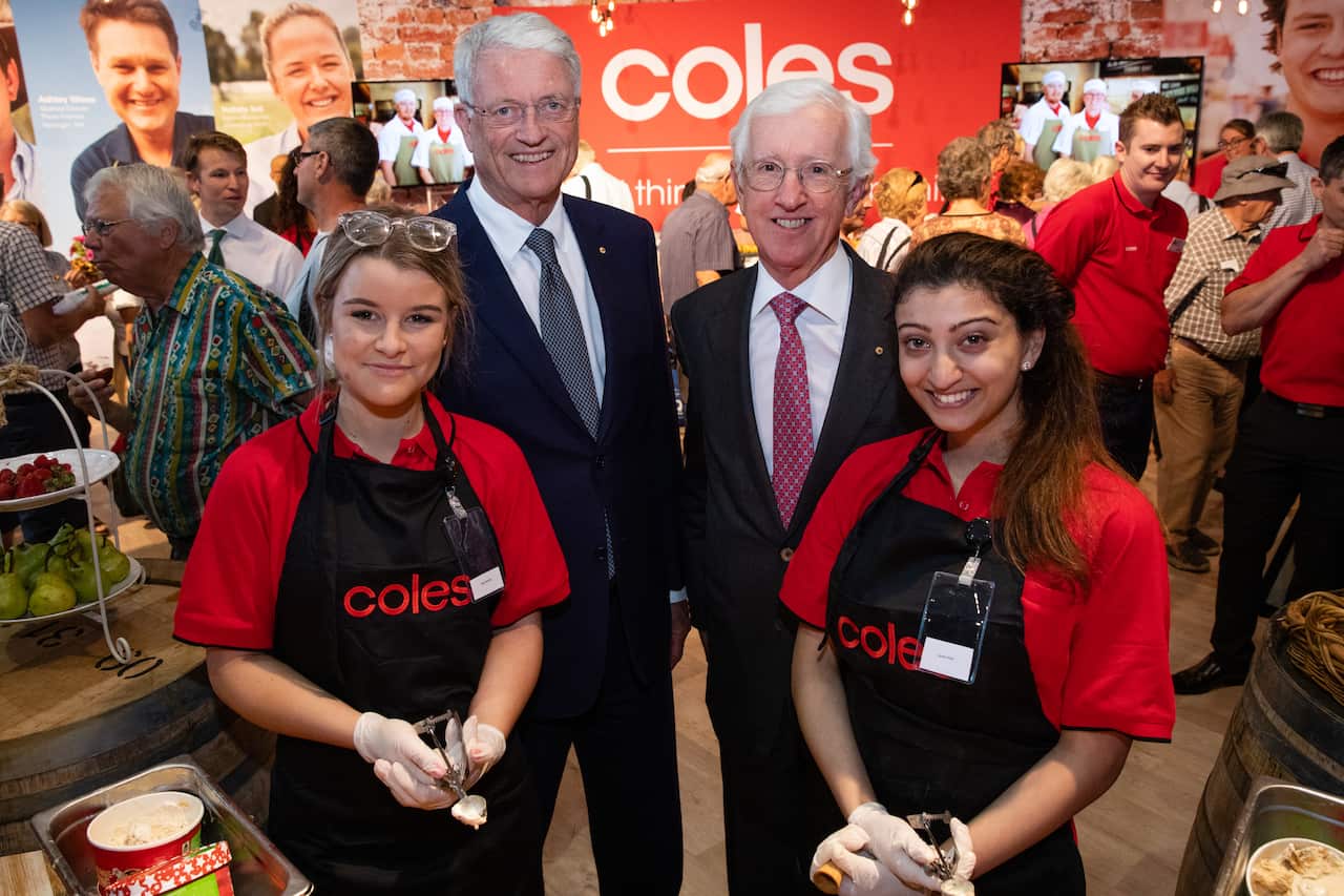 Two men wearing suits, ties and glasses stand smiling behind two women who are wearing red t-shirts and aprons that have "Coles" written on them.