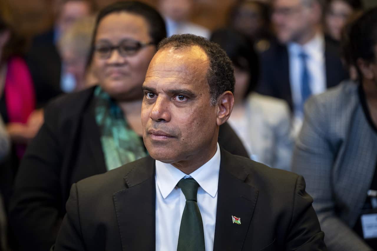 A man in a black suit sits in the audience at an event.