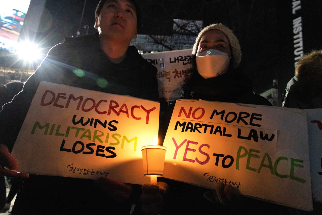 Two protestors hold signs in darkness lit by candlelight that say "Democracy wins militarism loses" and "No more martial law yes to peace" 