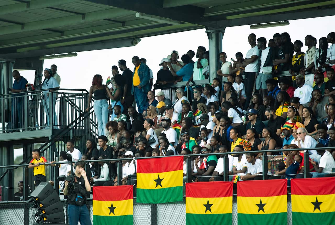 A large crowd of spectators fills a covered stadium grandstand decorated with multiple large flags of Ghana draped along the front railing.