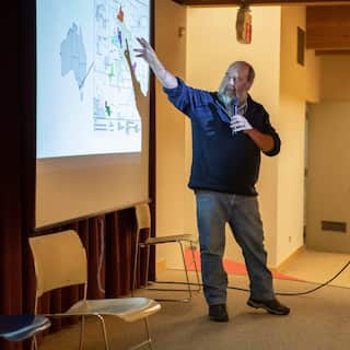 Denis Rose, a 71-year-old Gunditjmara man wearing a navy fleece and denim jeans, stands in front of a projector screen holding a microphone.