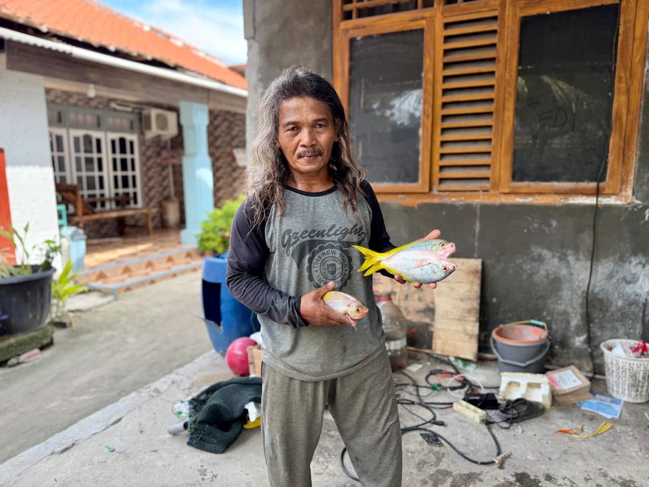 A man wearing light grey pants and a grey long-sleeved t-shirt with black sleeves holds up two small silver fish with yellow tails in his left hand. He is also holding a small fish in his right hand.