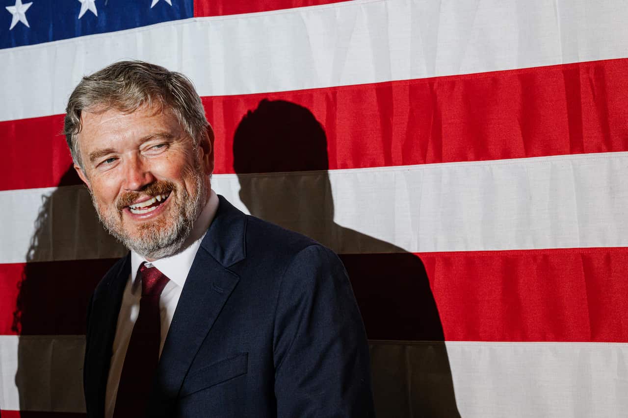 A smiling man in a suit in front of a US flag