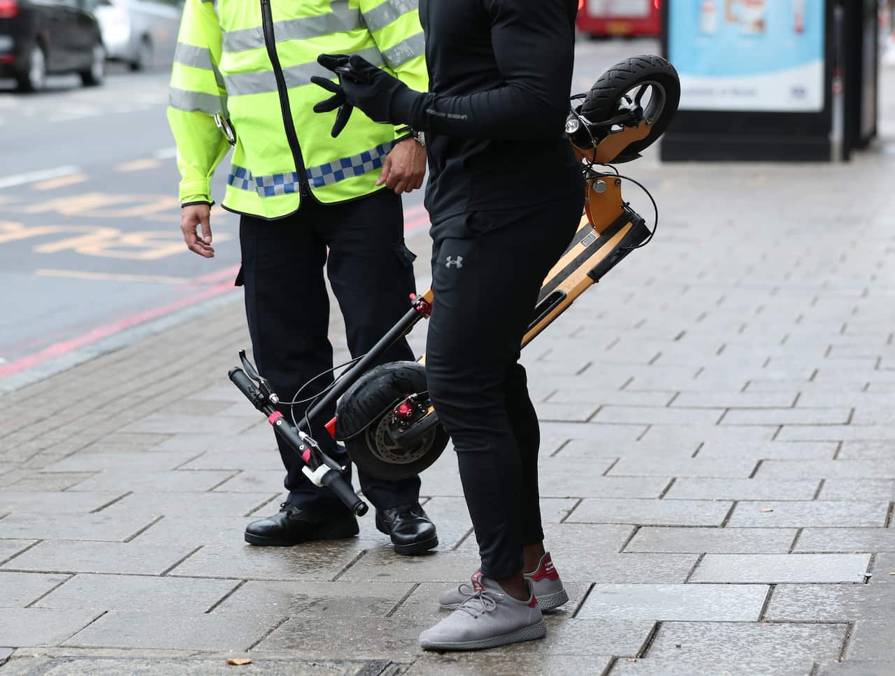 A person holding their e-scooter folded up, while standing beside a police officer.