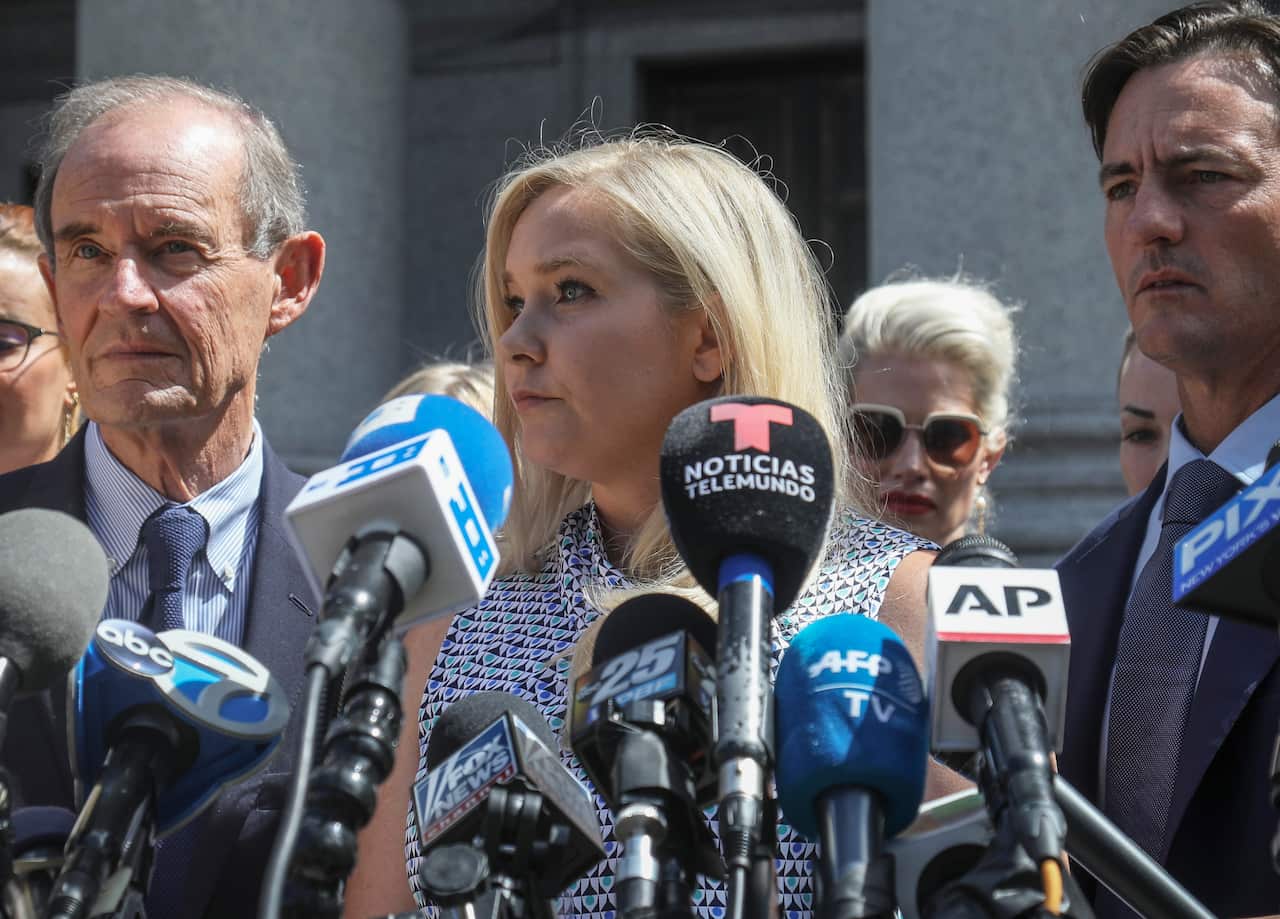 A woman with blonde hair is holding a news conference with people standing behind her.