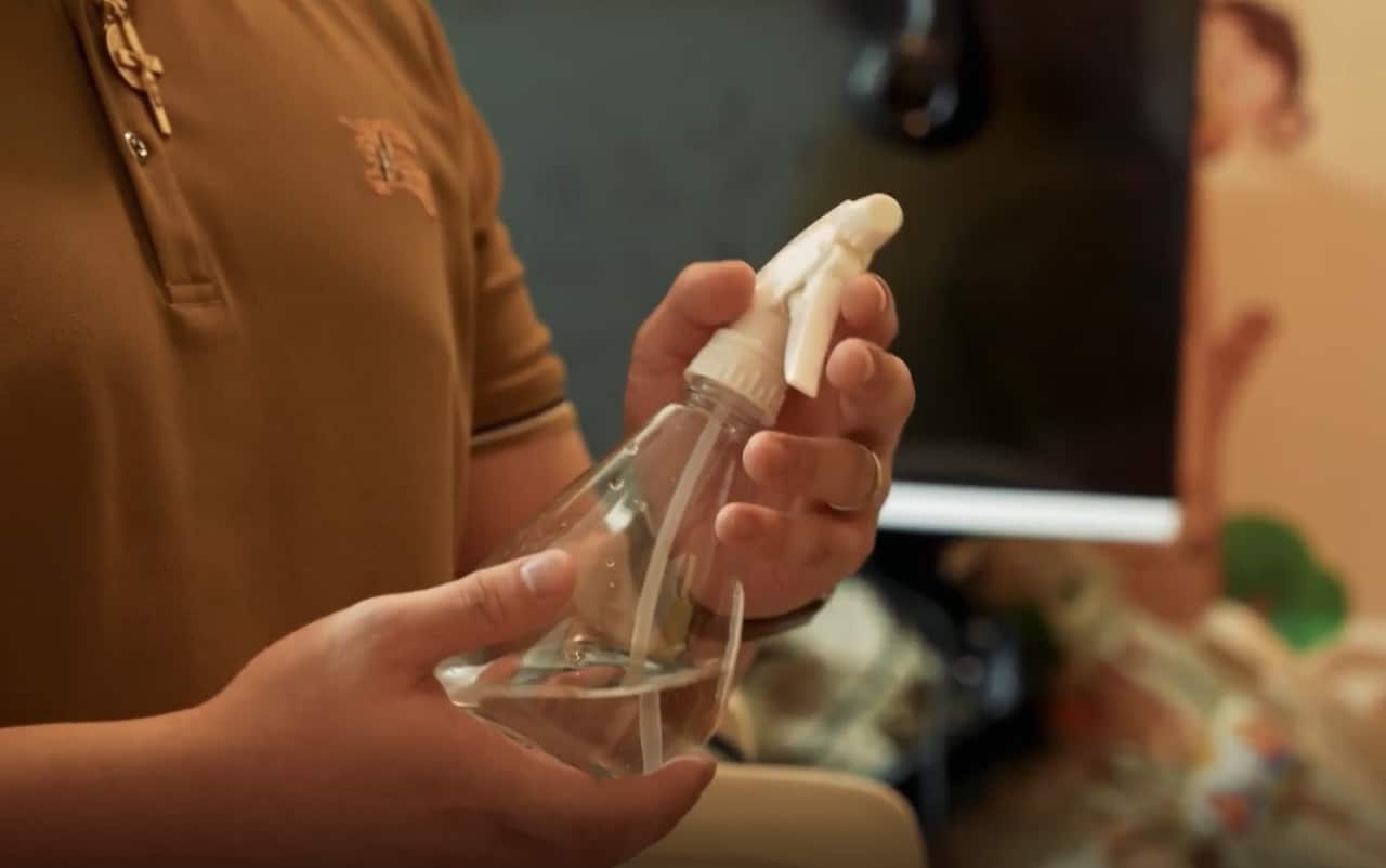 A man's chest and hands are visible. He is holding a clear spray bottle filled with liquid. 