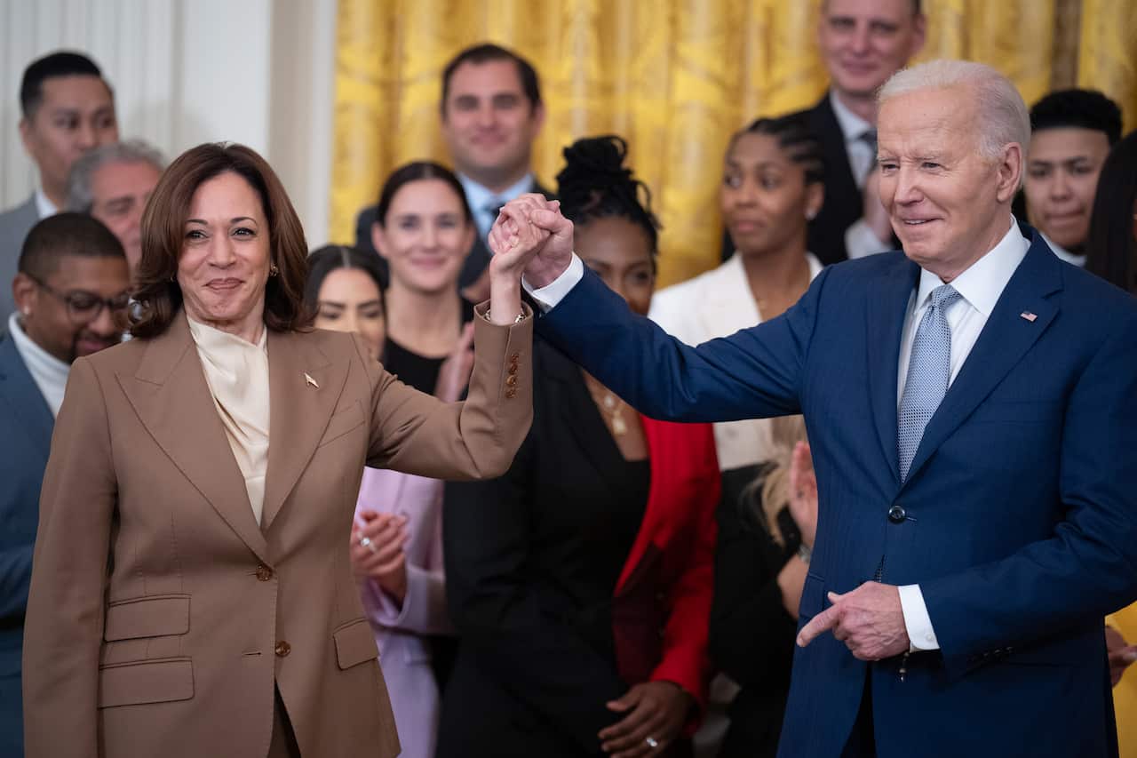 Kamala Harris (left) and Joe Biden (right) hold hands and raise their arms in celebration. A group of people stands behind them, clapping.