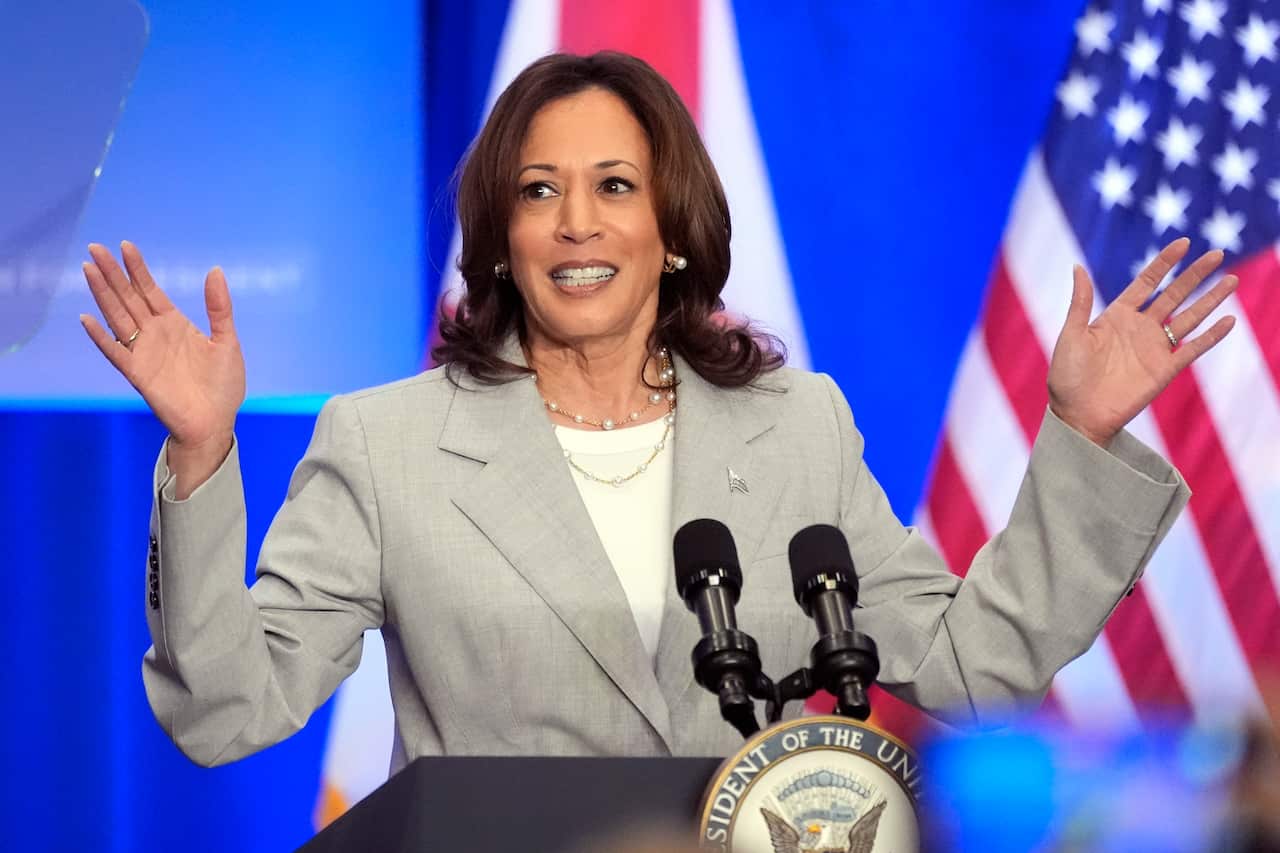 A woman stands in front of a lectern, gesturing with her hands. The American flag hands behind her