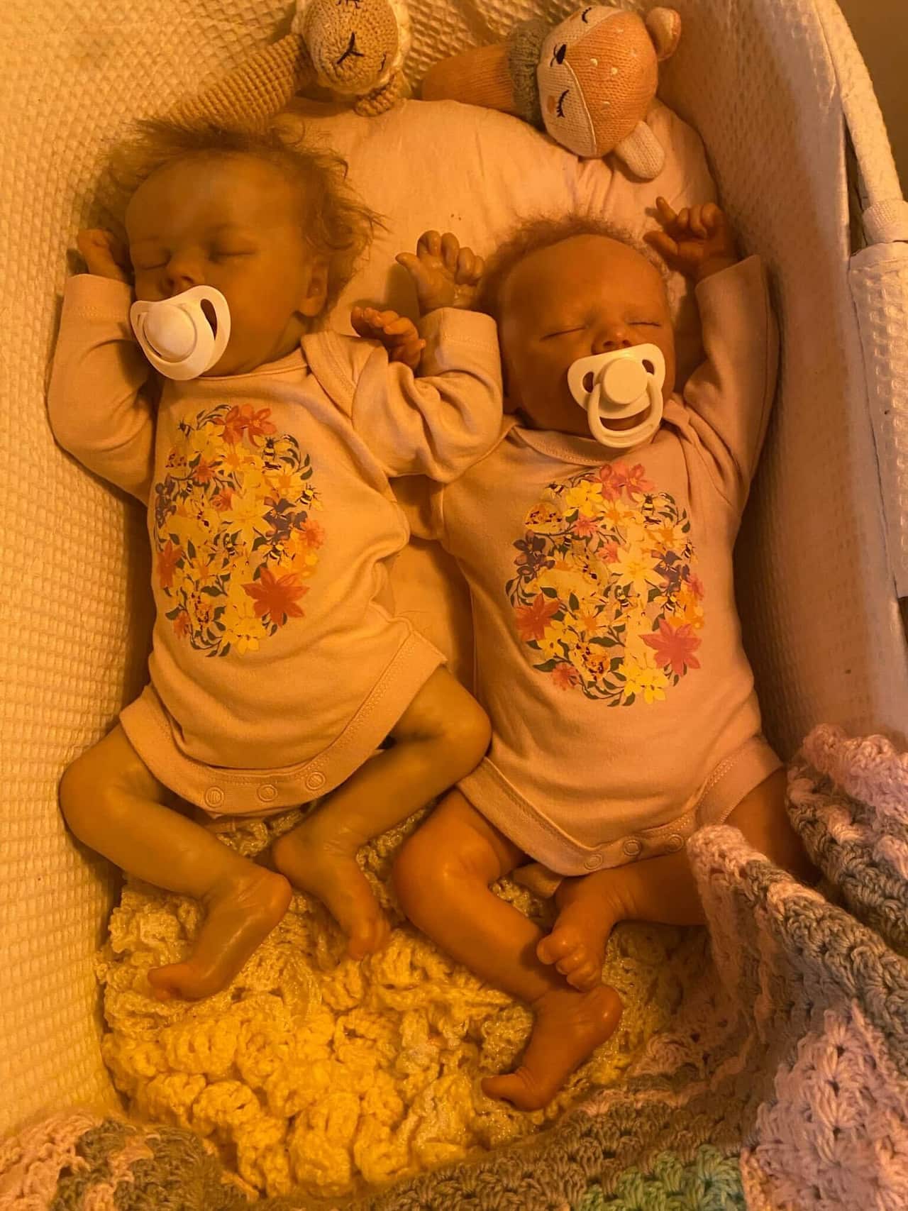 Two dolls resembling twin babies lie in a bassinet, complete with pacifiers, a blanket and stuffed animals