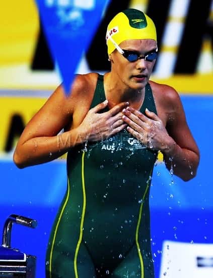 A swimming athlete stands in her swimsuit, swimming cap and googles, wet from the pool.