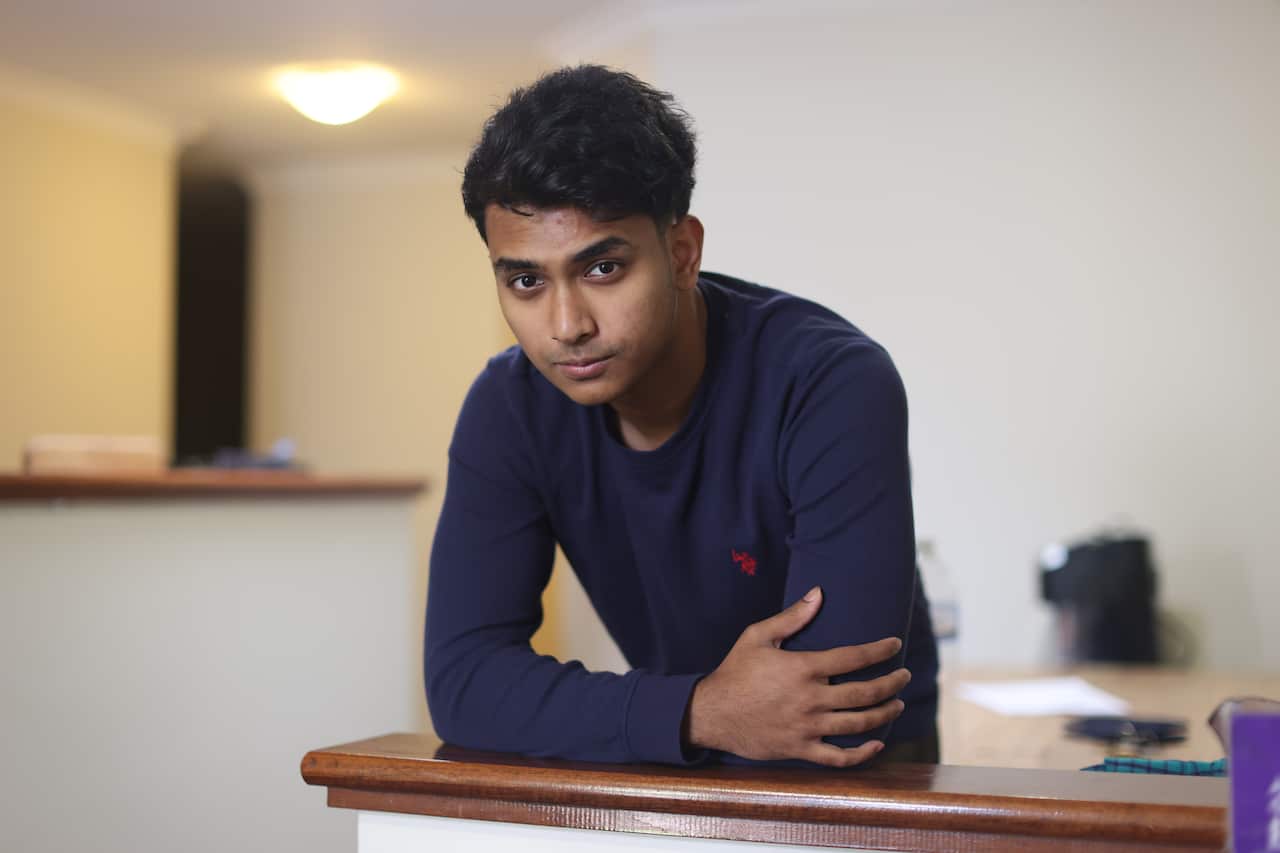 A young man leans on a wooden ledge, looking serious.