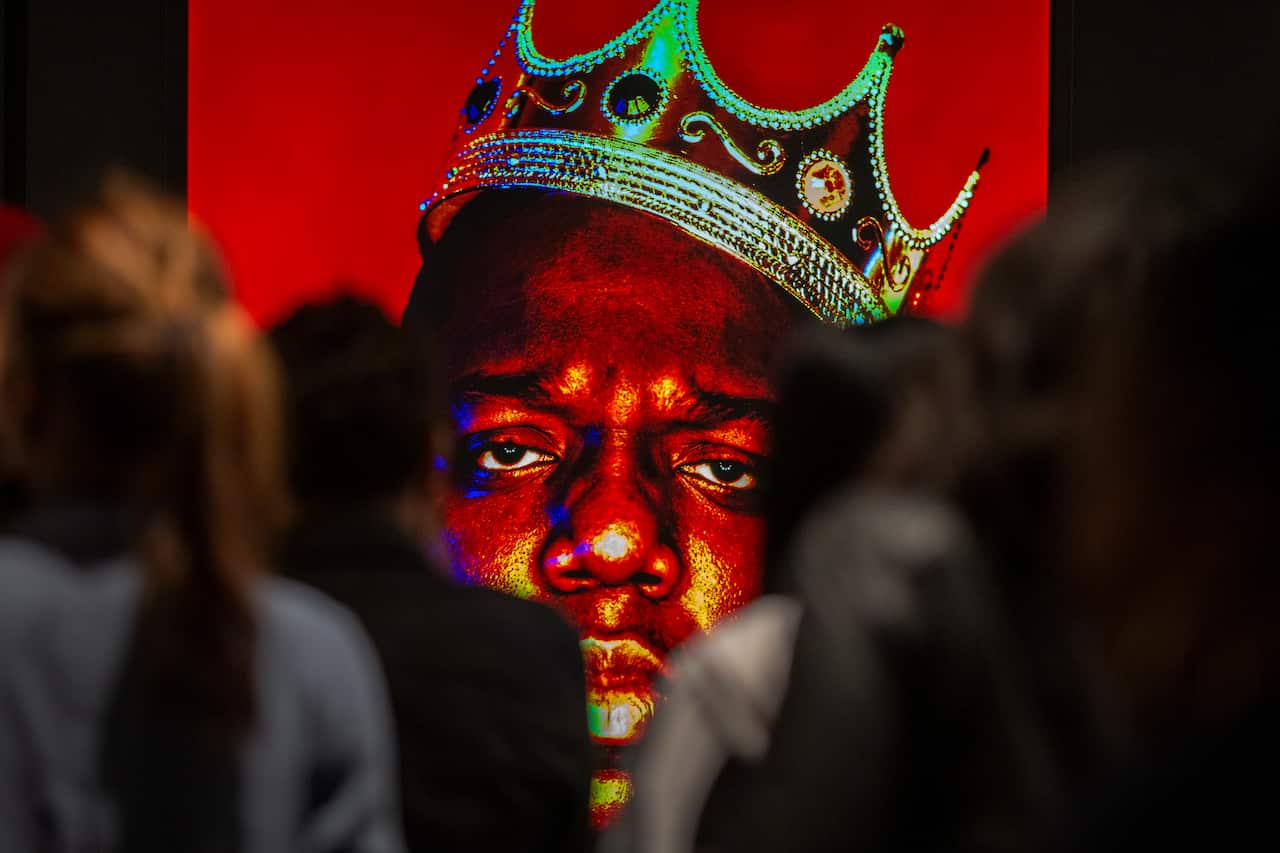 A crowd is looking at a photograph of US rapper Biggie Smalls wearing a crown. It's a close-up of the rapper's face taken against a red backdrop.
