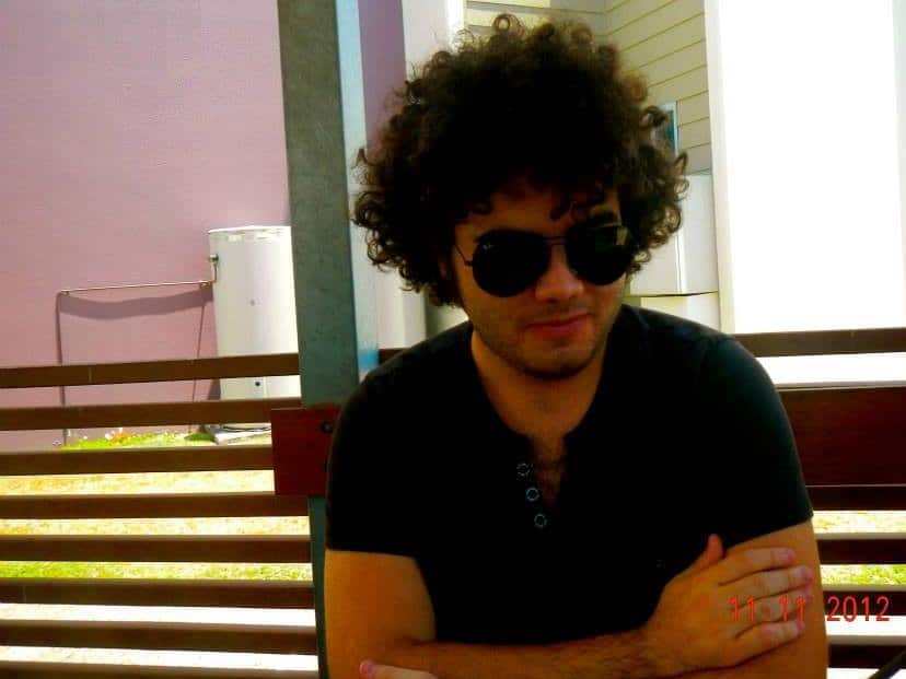 A man with dark curly hair and wearing sunglasses and black t-shirt sitting outside with his arms crossed.