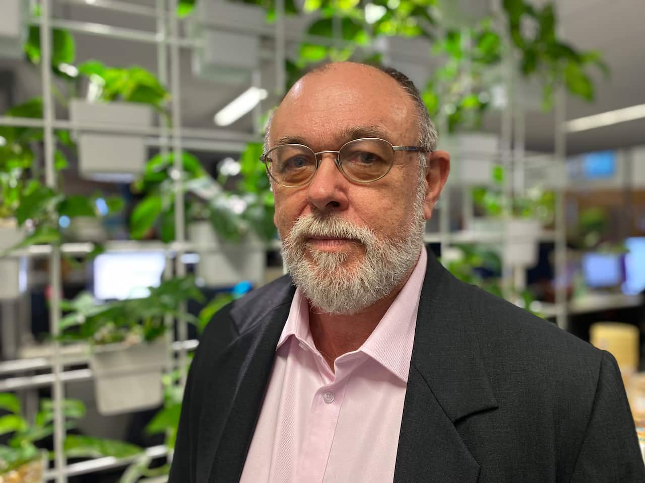 A man with a beard and glasses and wearing a pink shirt and dark suit jacket standing inside