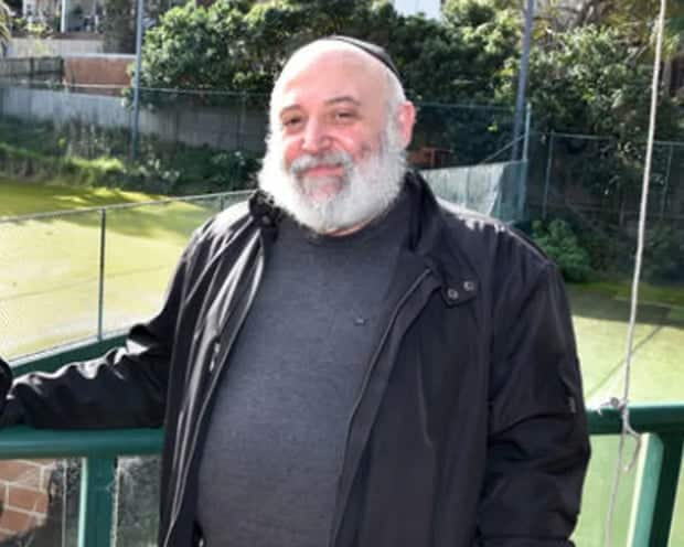 An elderly man wearing a black jumper and jacket is standing in a balcony.
