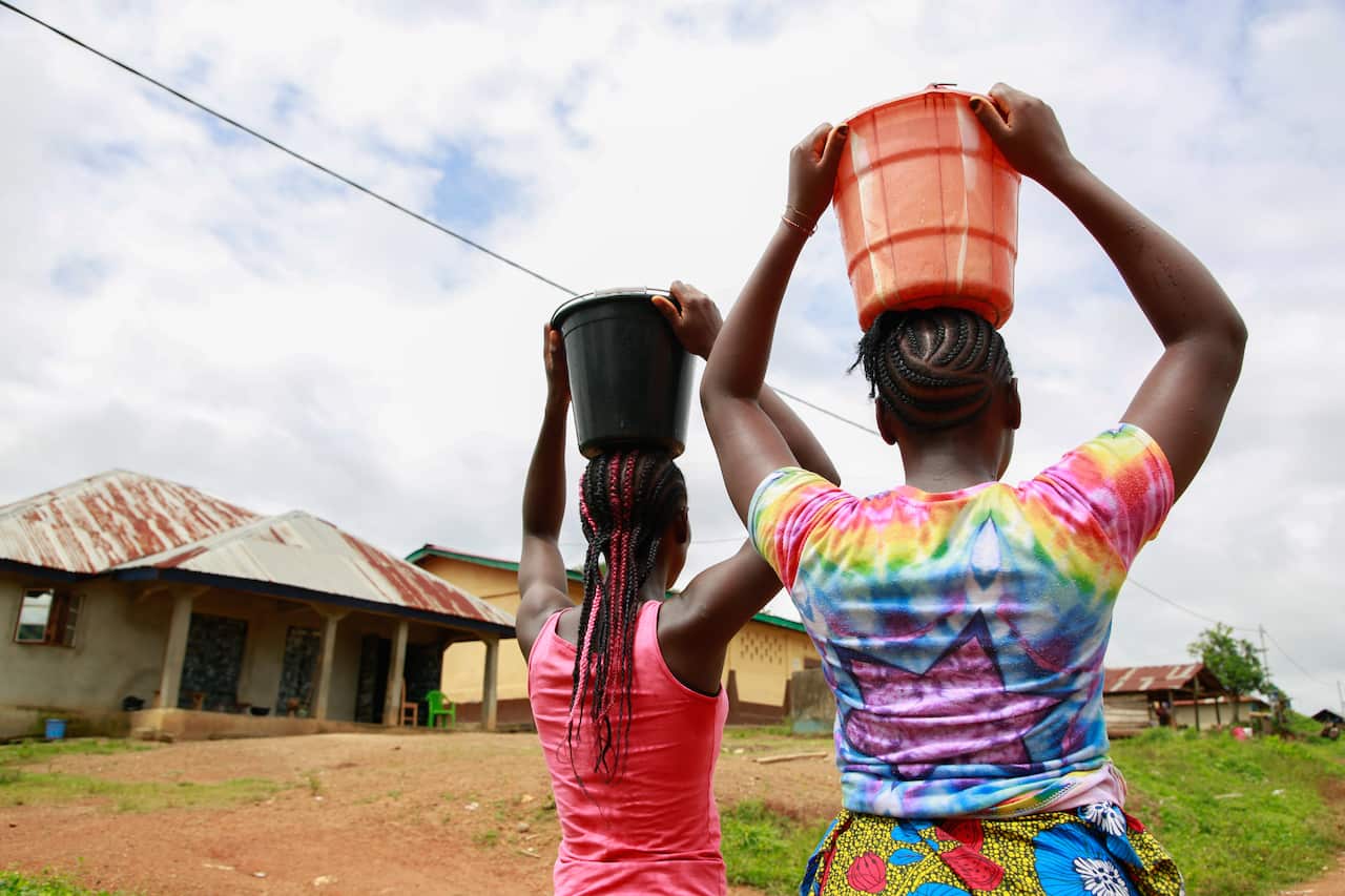 Two teenage girls, one wearing a pink tank top and the other a tie dyed t-shirt, carry buckets full of water on their heads.