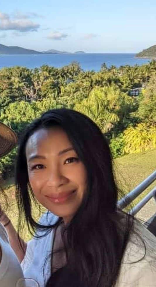 A woman with long dark hair posing for a photograph. There is bushland in the foreground and a body of water in the background