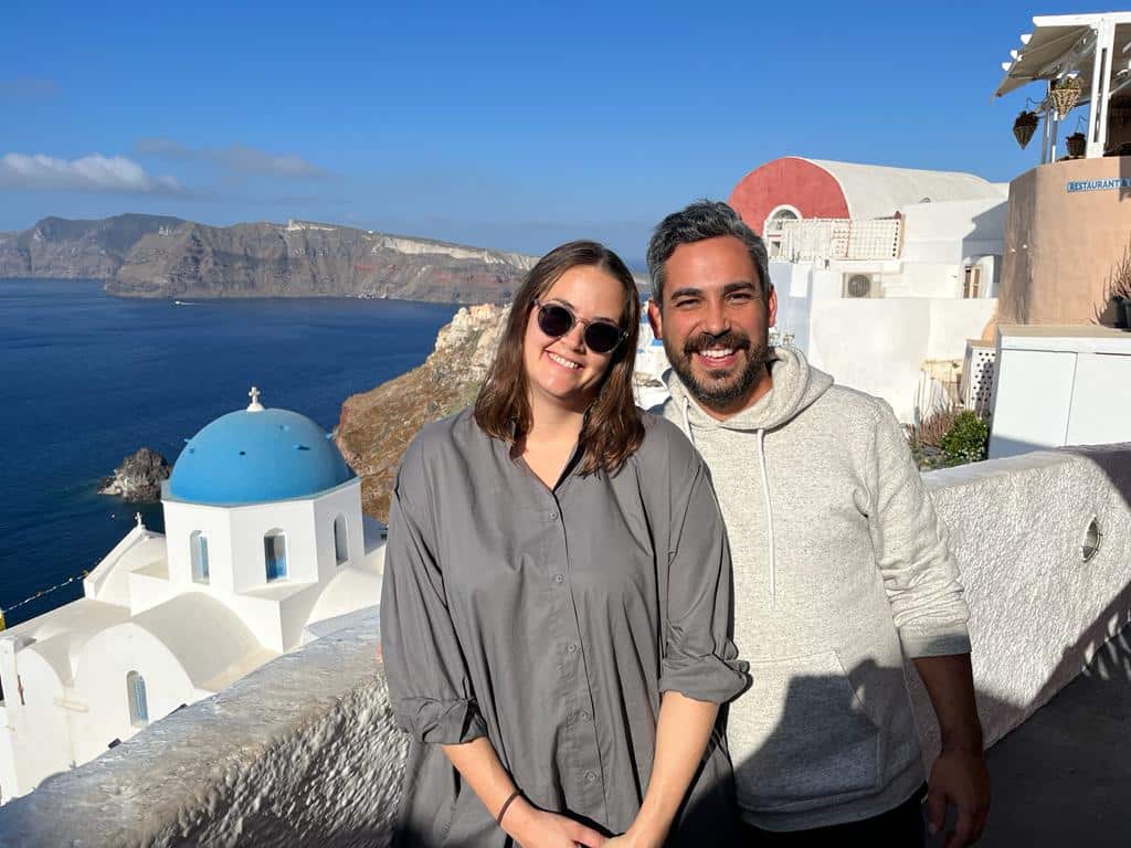 A man and a woman standing in a seaside village in Greece. There is a white church with a blue roof and the sea in the background and cliffs in the distance