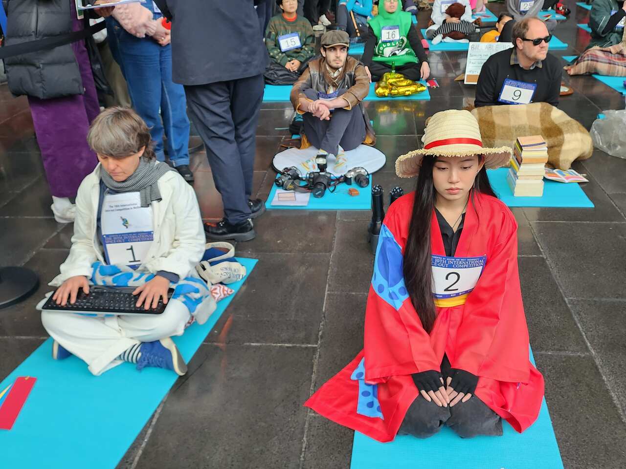 A person holding a keyboard sits on a blue mat next to a woman in traditional red Japanese dress.