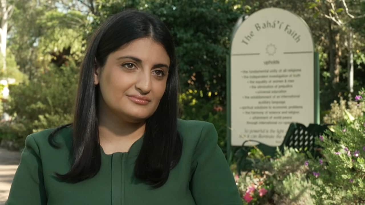 A woman with dark hair, wearing an olive green top. She is in a garden.