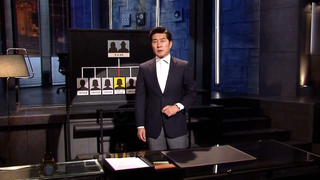 A still from a TV program showing a middle-aged Asian man in a white shirt and dark suit standing in a TV studio. 