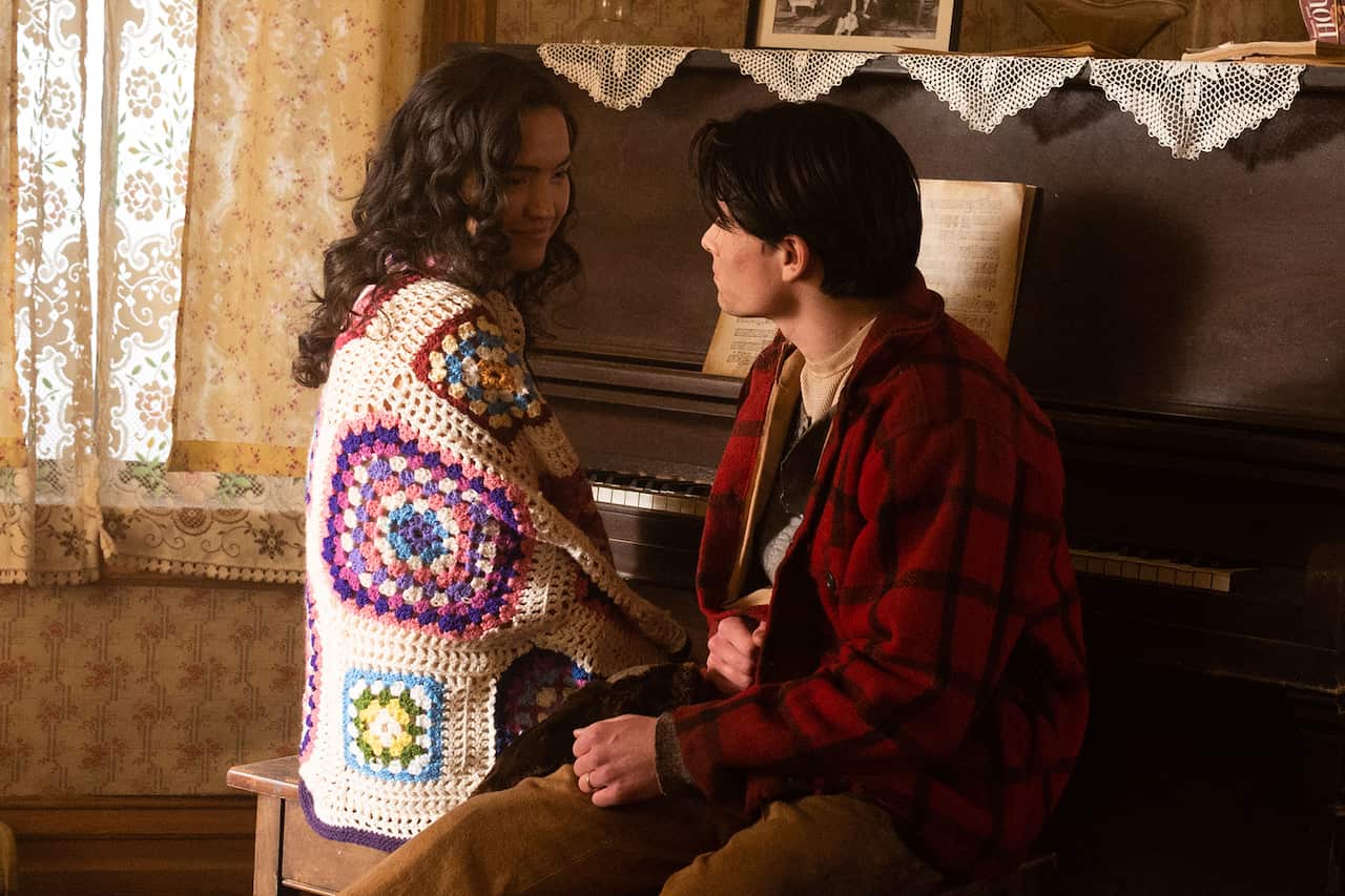 A young woman with long, curly dark hair and wrapped in a colourful crocheted blanket, sits at a piano, looking sideways and smiling at a young man in a red jacket, who sits on the piano stool beside her.
