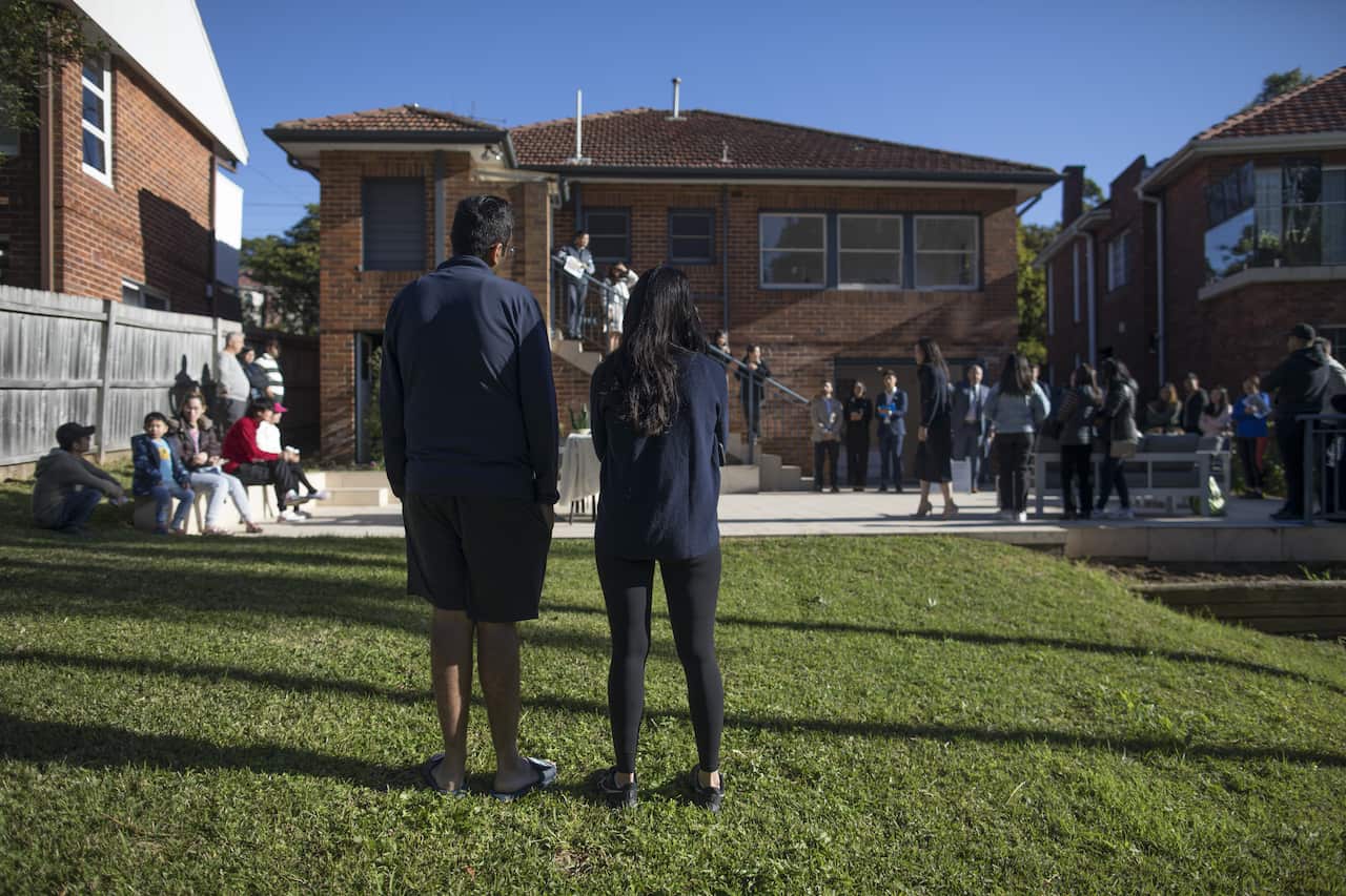 A man and a woman wearing navy jackets, standing on a lawn out the front of a house. People are gathered near the house.