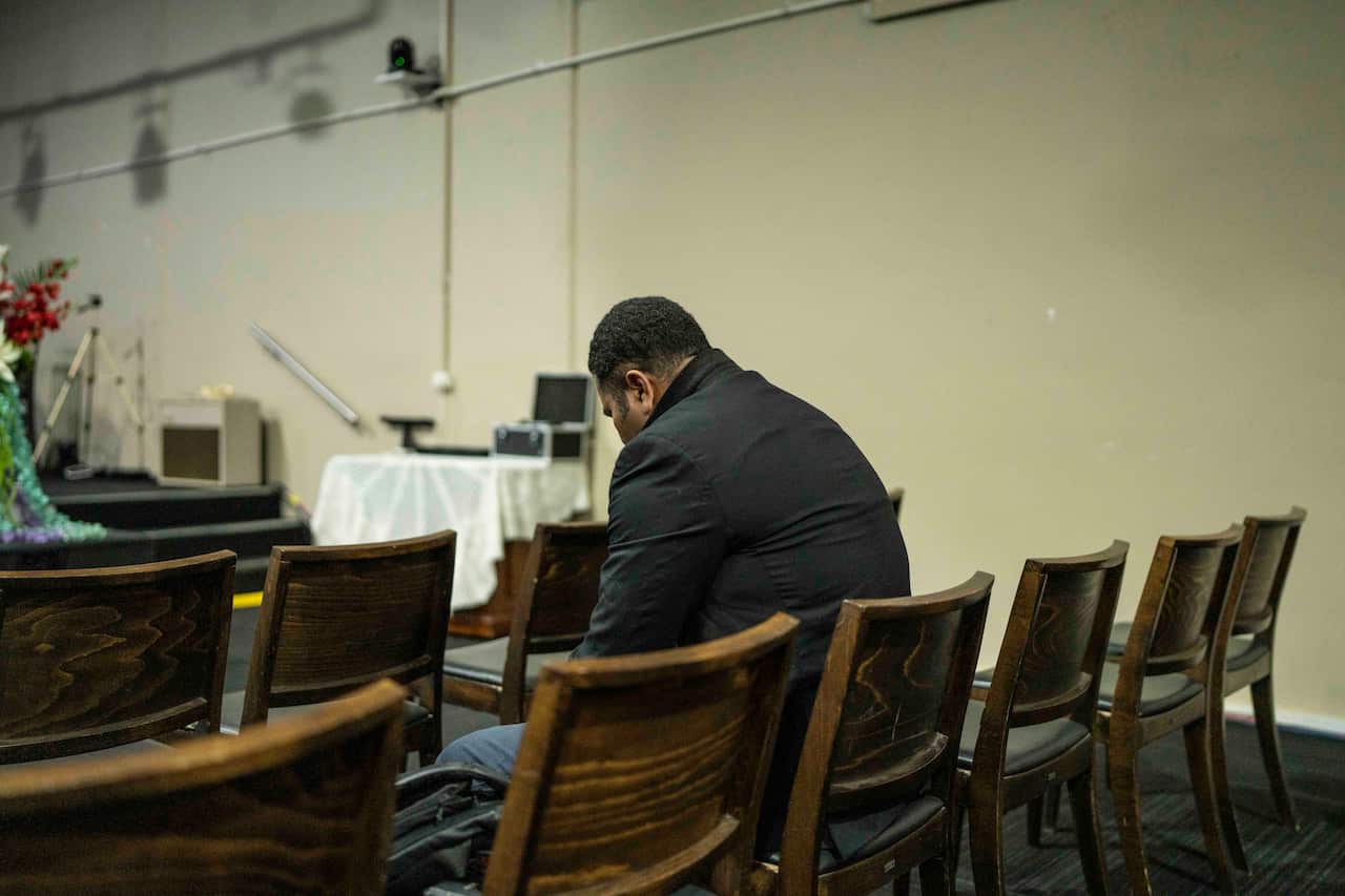 A Pasifika man wearing a dark jacket sits on a wooden chair in a community church, facing away from the camera.