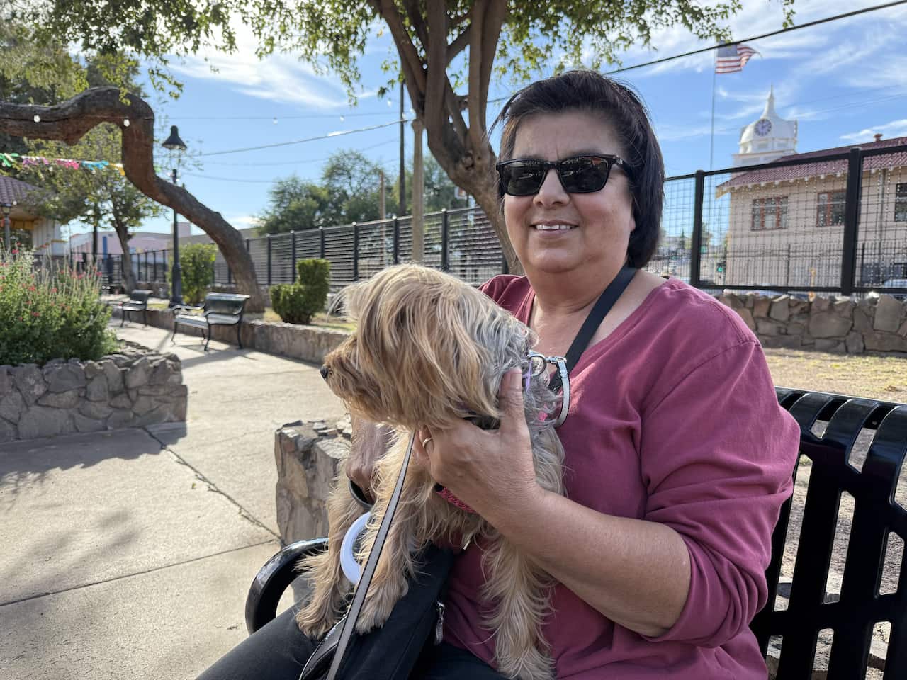A woman sitting on a park bench with her small dog, an American flag flies in the background.