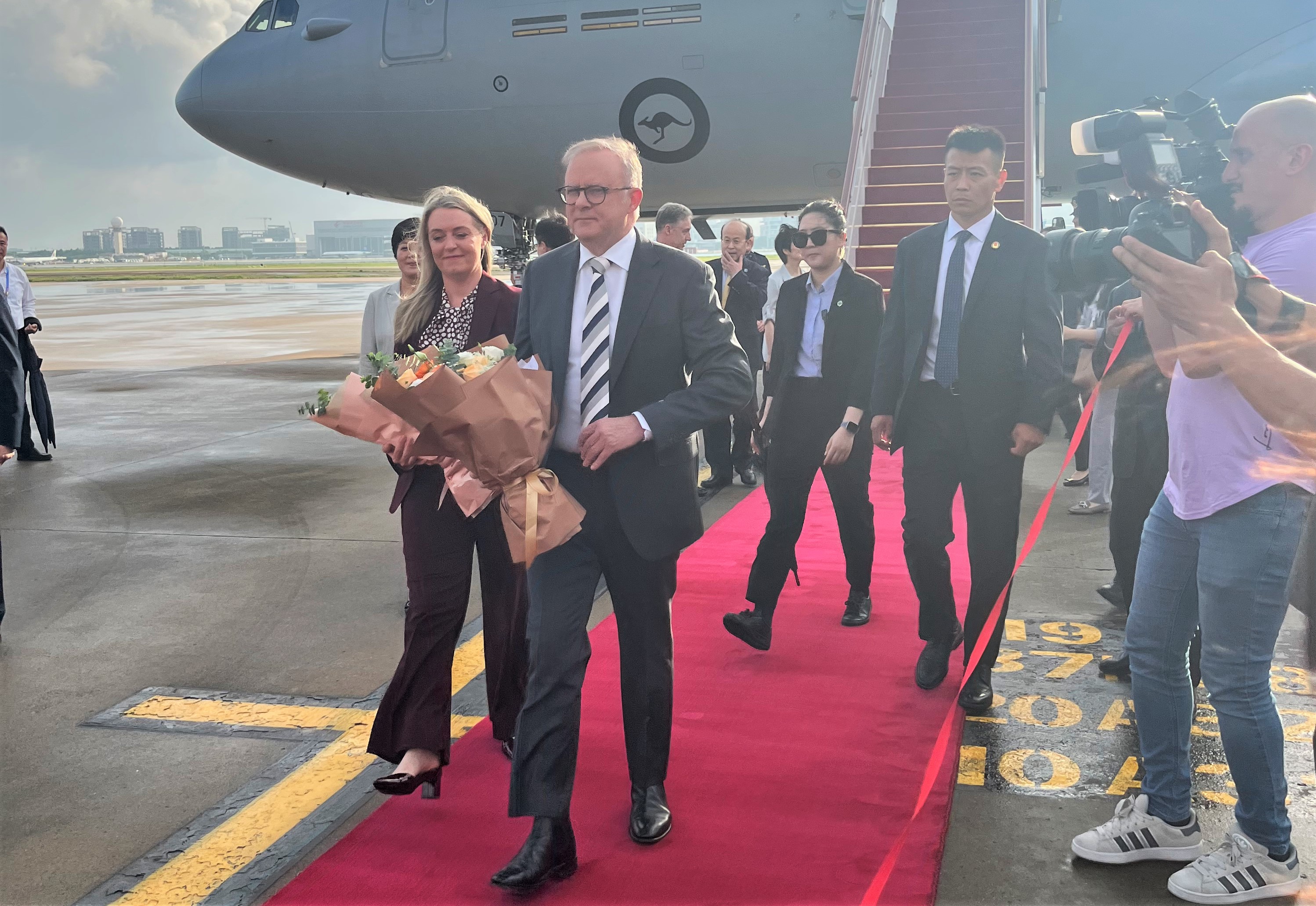 Anthony Albanese, a middle aged man with glasses, wearing a dark suit, stripes grey, black and white tie, holding a bunch of flowers, walking down a red carpet off a large airplane, surrounded by people.
