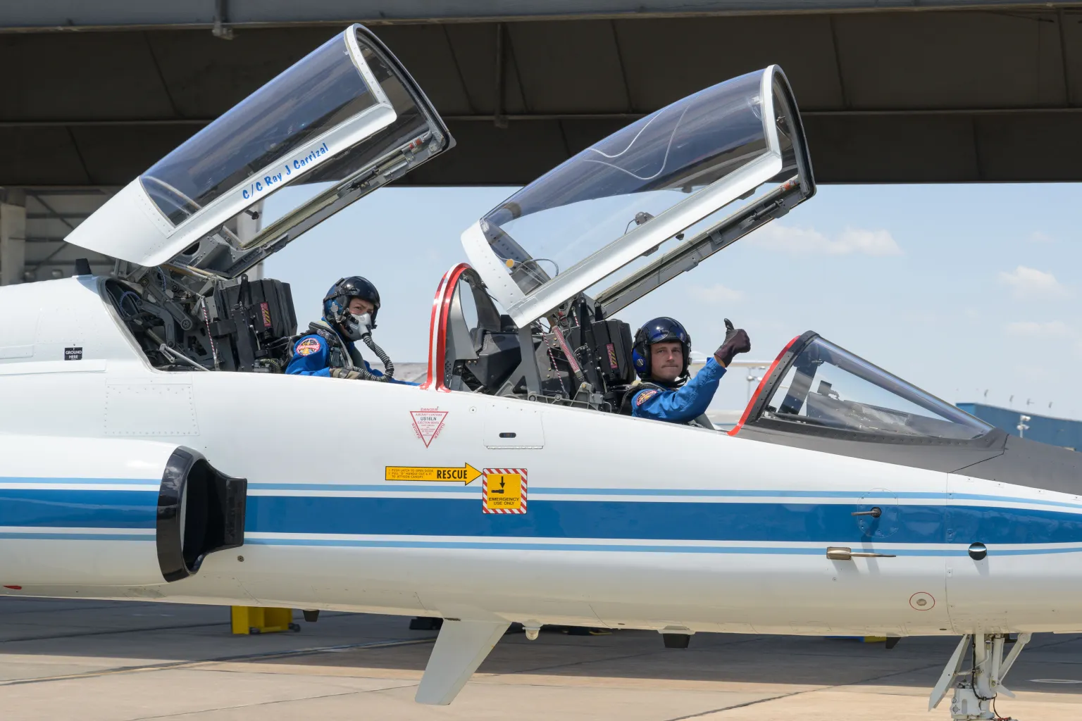 Two men on a T-38F flight jet, wearing helmet, with one giving a thumb up.