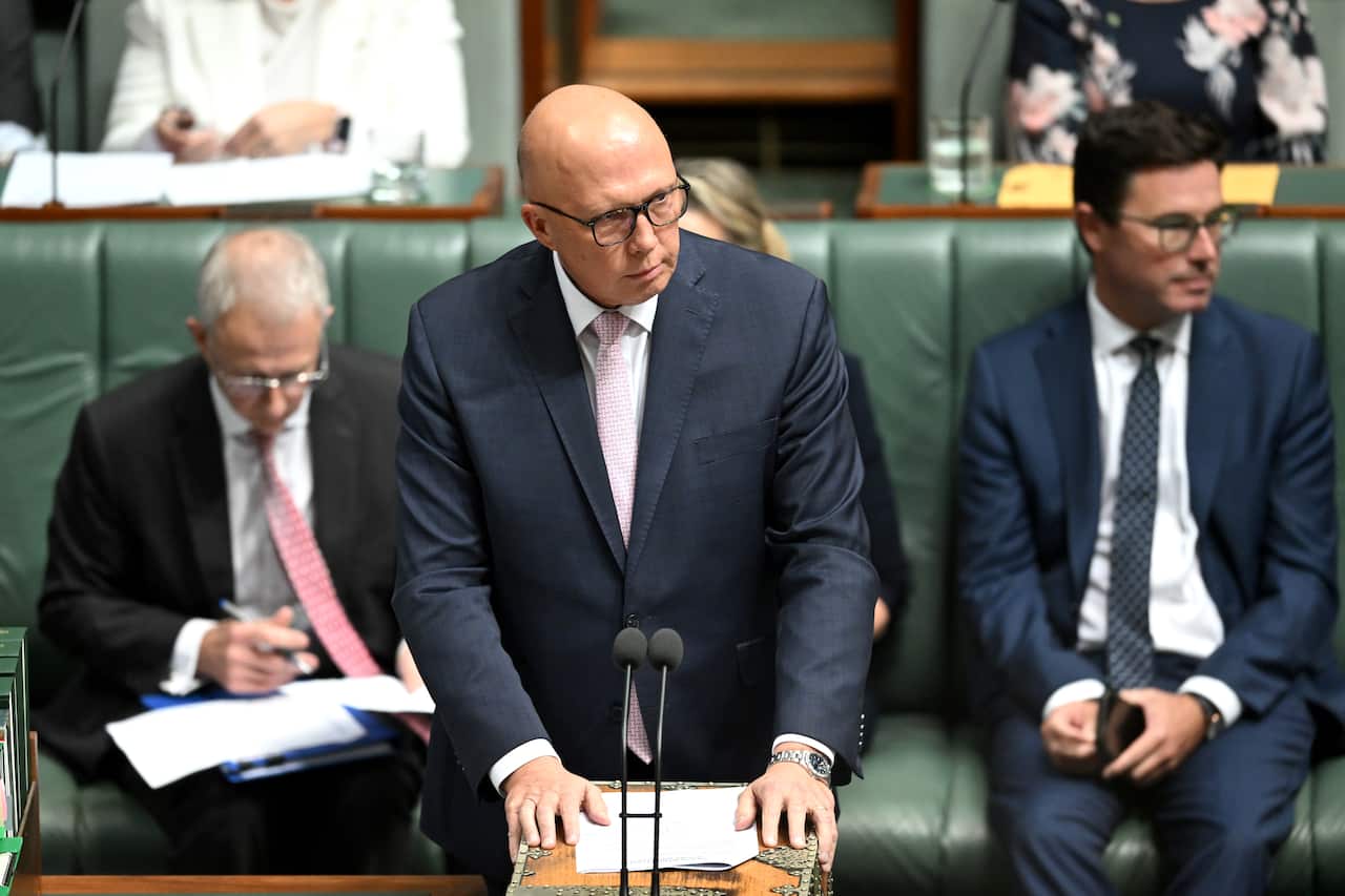 A bald man wearing glasses and a black suit stands in front of a microphone in parliament.