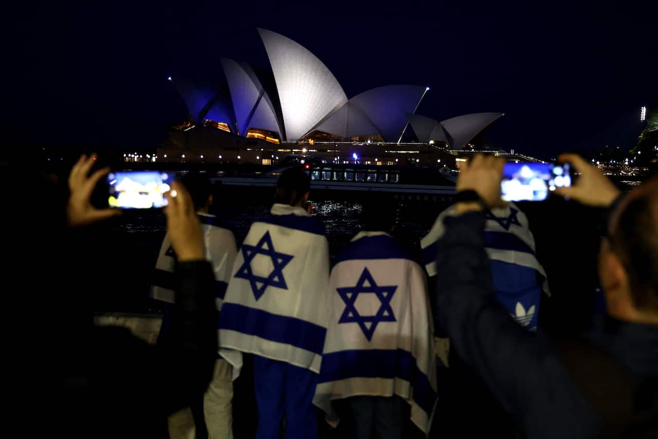 People with Israeli flags watch the Opera House while it is illuminated in blue to show solidarity with Israel.
