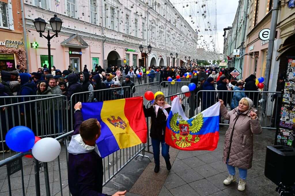 Three people standing on a footpath hold a Russian and a Moldovan flag in front of a group of people standing on a street. 