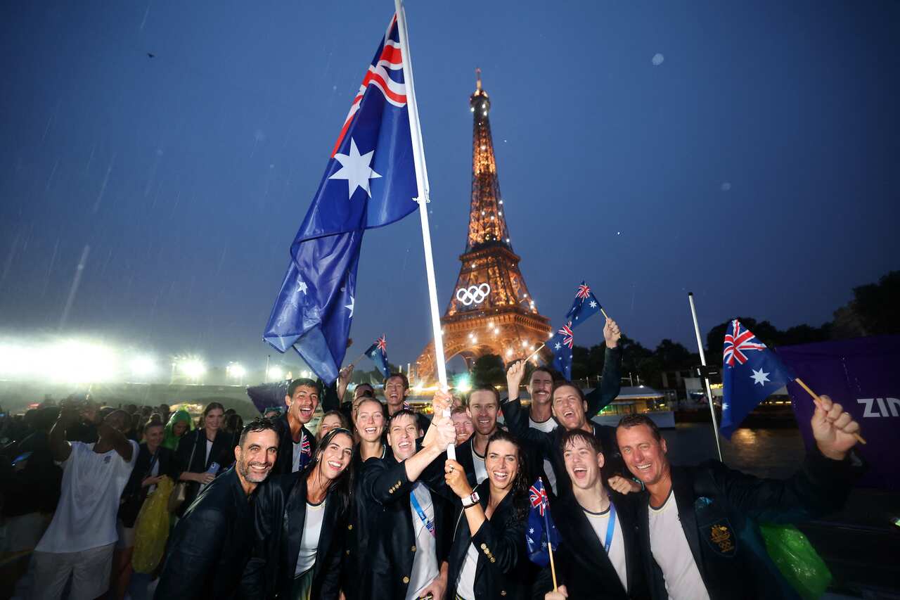 Some of the Australian Olympic team at the Paris Games' opening ceremony. They are smiling and cheering, and some are waving Australian flags. The Eiffel Tower is lit up in the background.