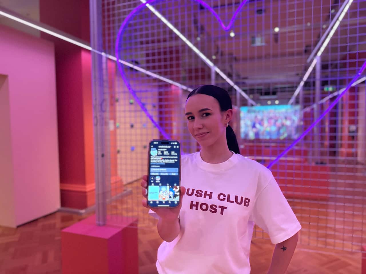 A woman in a white shirt holding her phone with an Instagram page at the camera. The room around her is tinted pink.