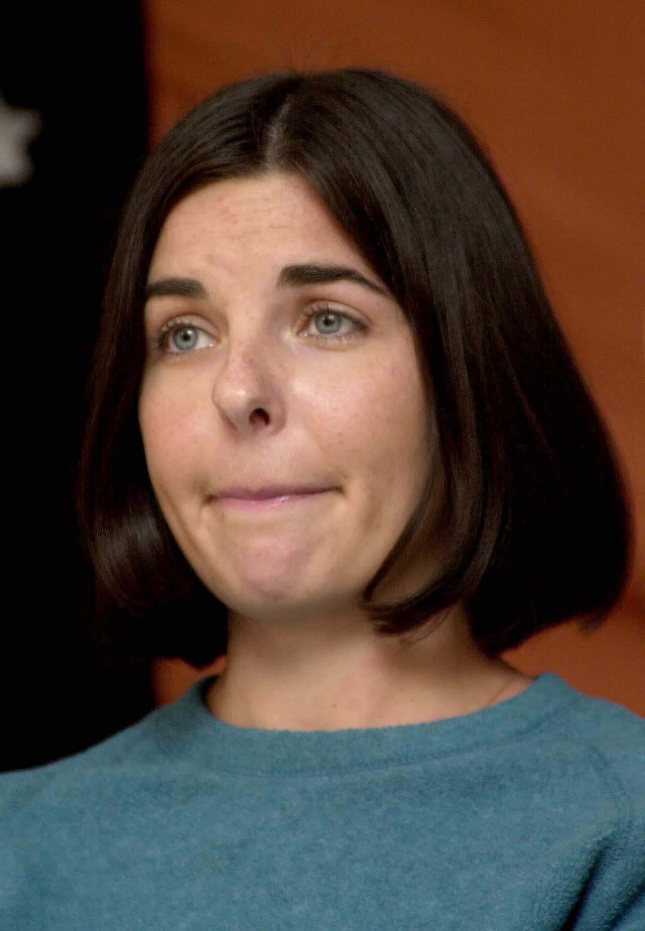 A woman in a green top and bobbed dark hair looks at the camera with a serious expression on her face.