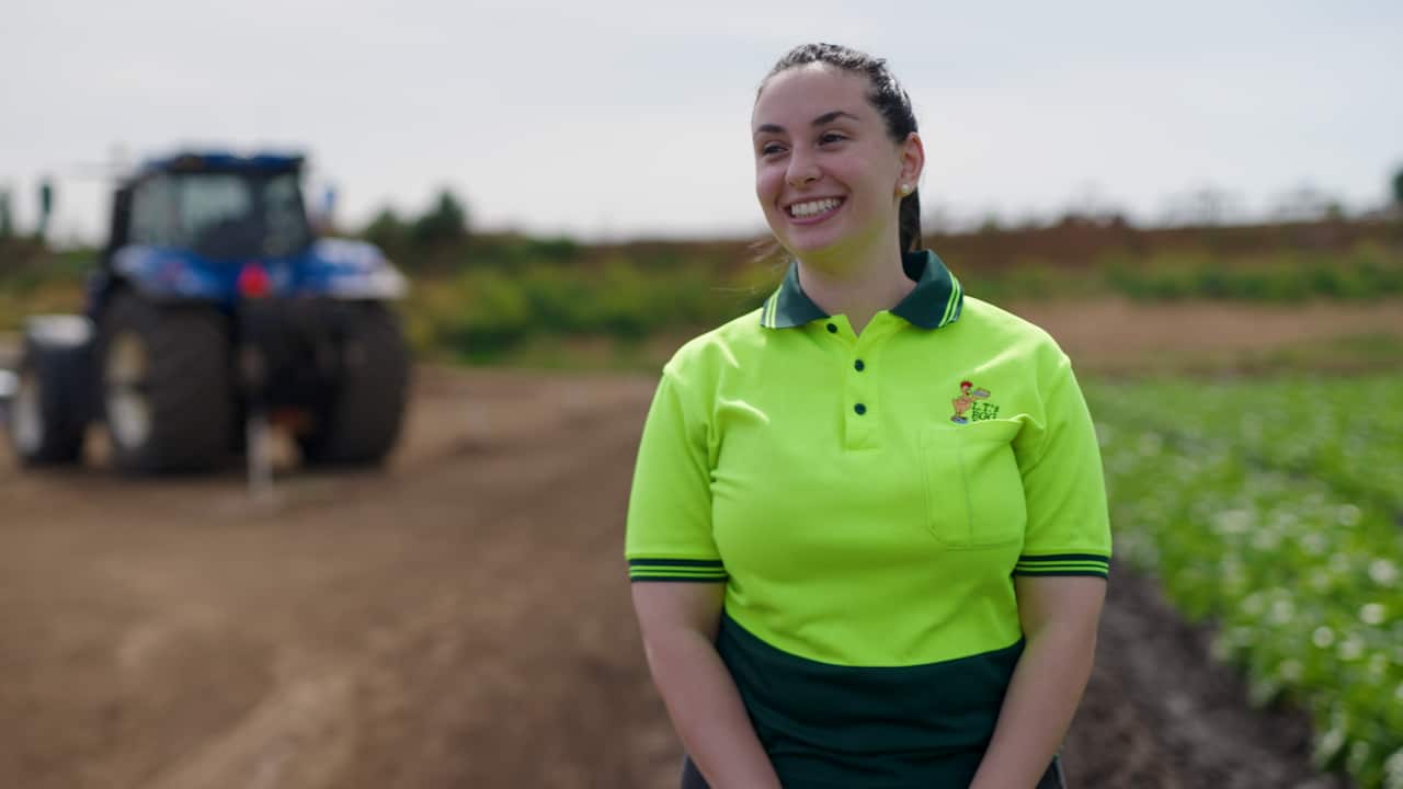 A smiling woman in a high-vis shirt stares into the distance, with vegetable fields and a tractor behind her
