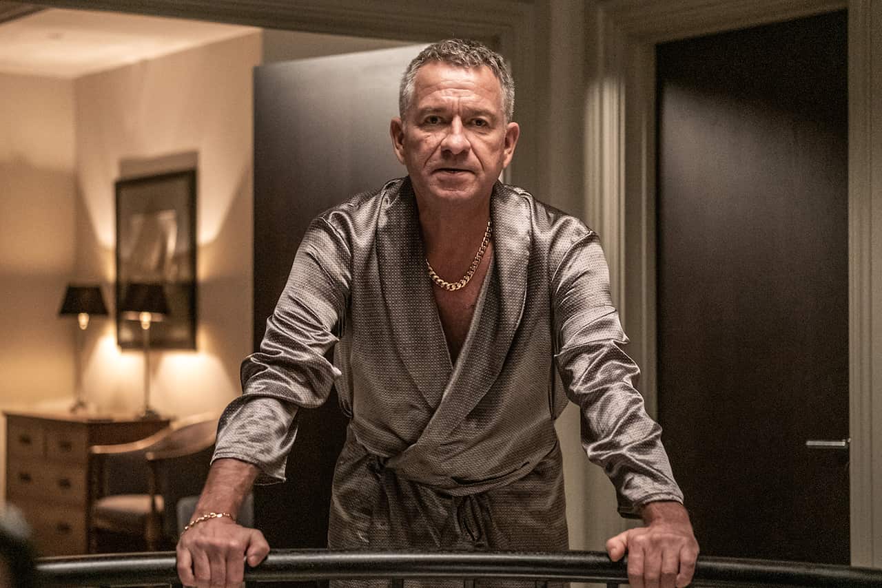 A man in a silky robe, with a chunky gold link necklace, stands on a balcony, leaning on the railing. A lamp-lit room can be seen through the door behind him.