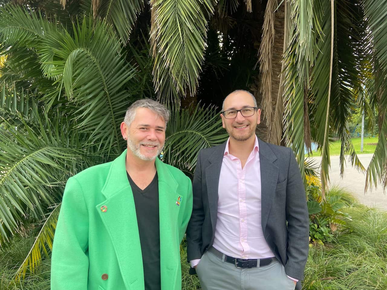 A man in a green blazer and a man in a pink shirt and grey blazer smiling standing in front of palm trees.