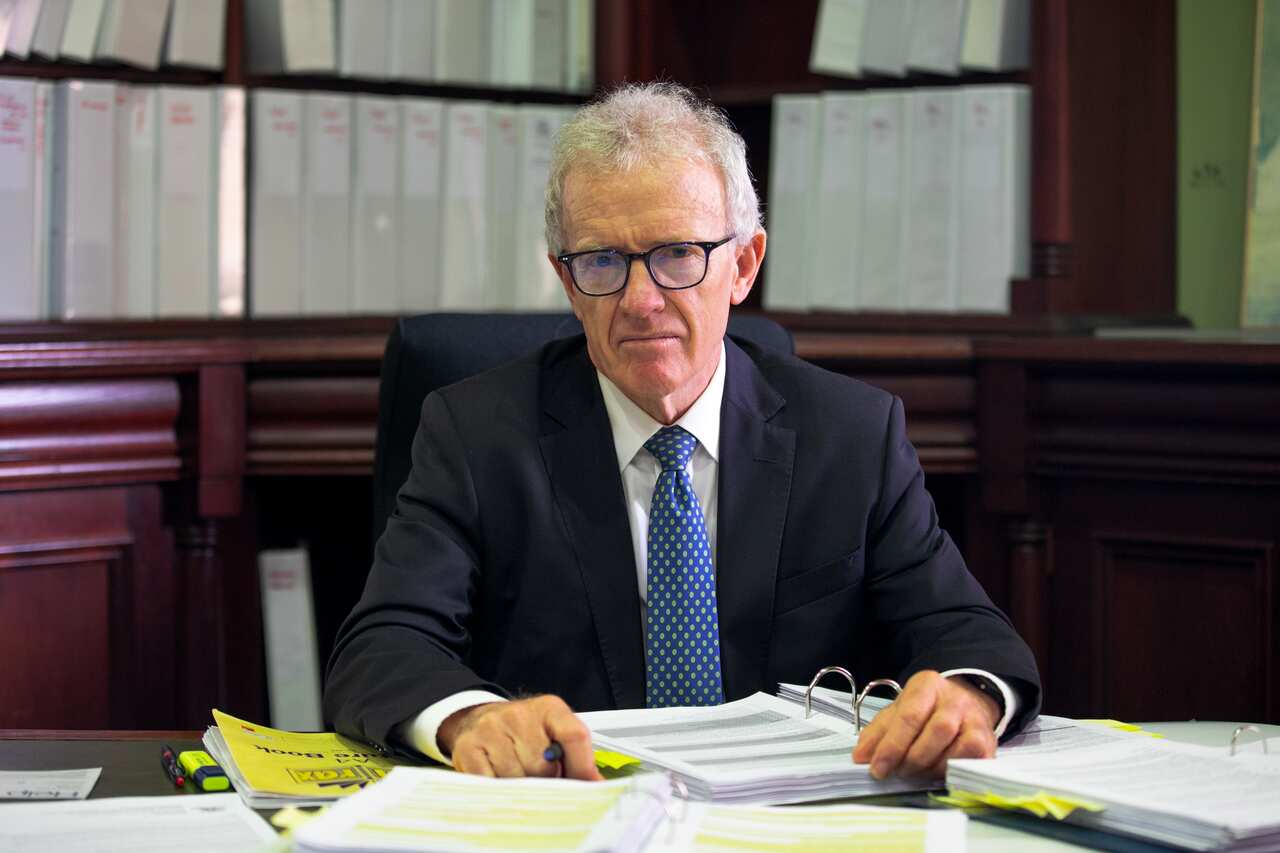 A man in a suit seated at a desk, with binders behind him.