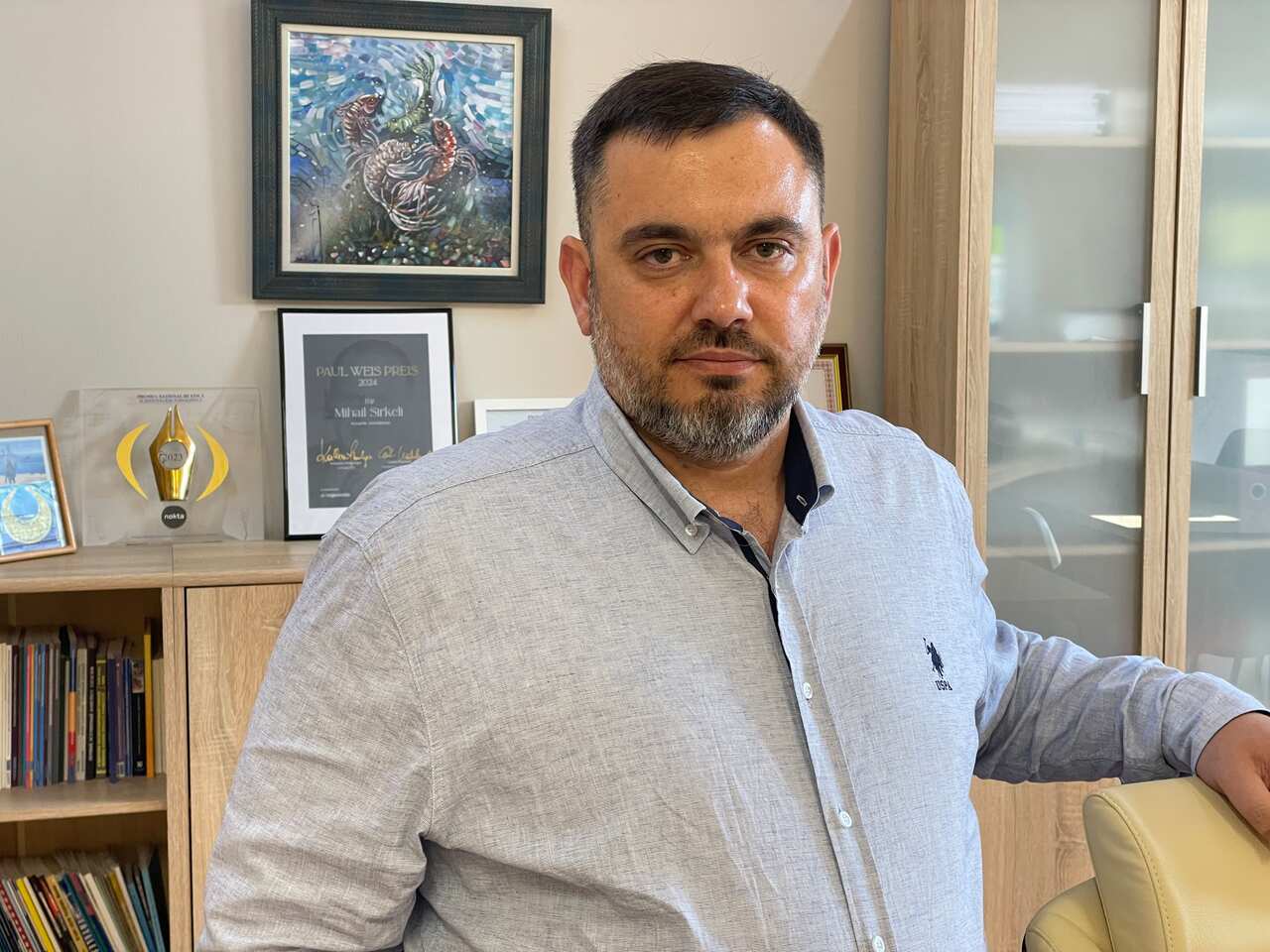 A middle-aged male with a short, greying beard wearing a light coloured buttoned shirt. 