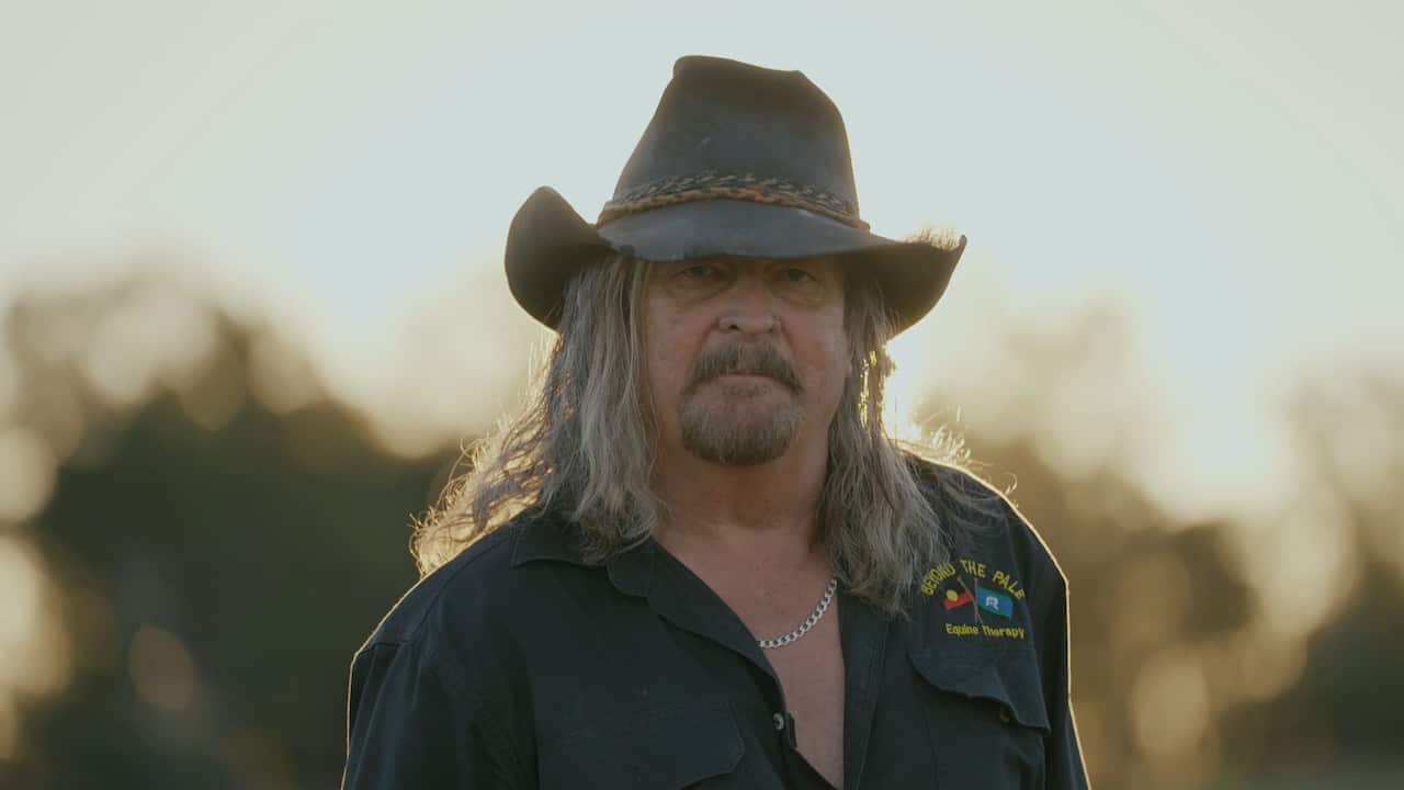 A portrait of a man with shaggy long grey hair, a goatee, a cowboy hat, a necklace and a shirt that reads "Beyond the Pale, Equine therapy"