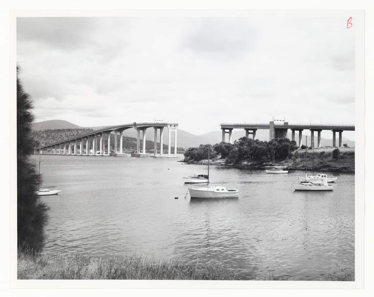 A black and white photo shows a large concrete bridge over a body of water missing a section in the middle. 