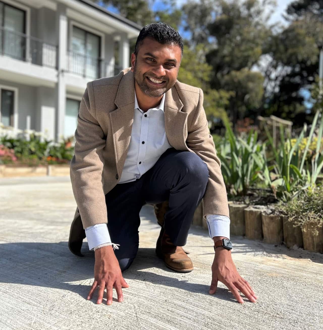 A man in a brown jacket crouches with his hands on a concrete path.