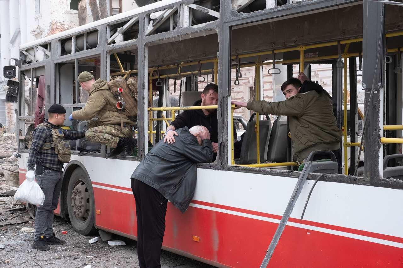 A man leans into the window of a red and white painted bus and holds his head in his hands. He is comforted by a man in the bus. There is another man in the bus to his right and two soldiers in uniform towards the front of the bus, which has no glass in its window frames.