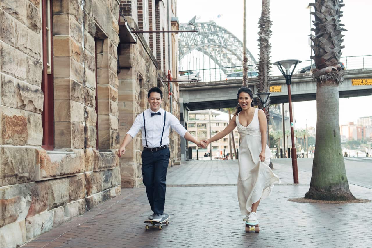 A couple holds hands while riding skateboards on a street.