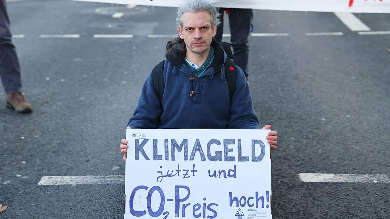 A man with grey hair wearing a blue hoodie and a backpack is kneeling on the ground holding a poster in the German language with both hands