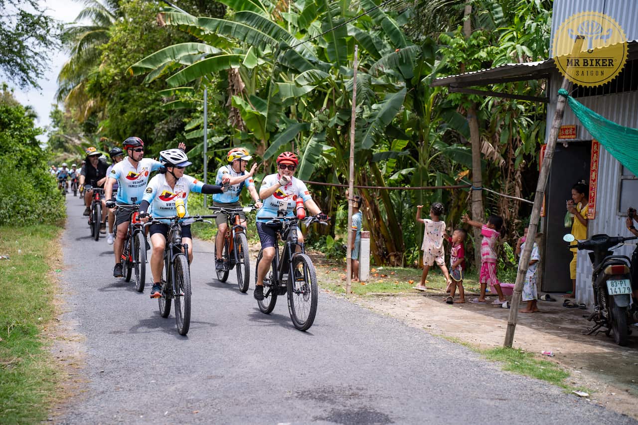 A group of people riding bicycles along a road surrounded by tropical green leafy jungle, led by four people wearing blue and white cycling outfits and helmets, who wave to some children standing outside a hut on the side of the road.